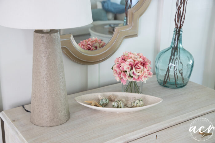 top of dresser with seashells in dish, pink flowers, blue vase and new lamp