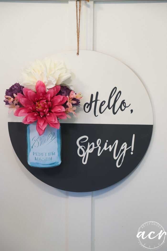 black and white round sign with hello spring, blue jar and pink colorful flowers hanging on wall